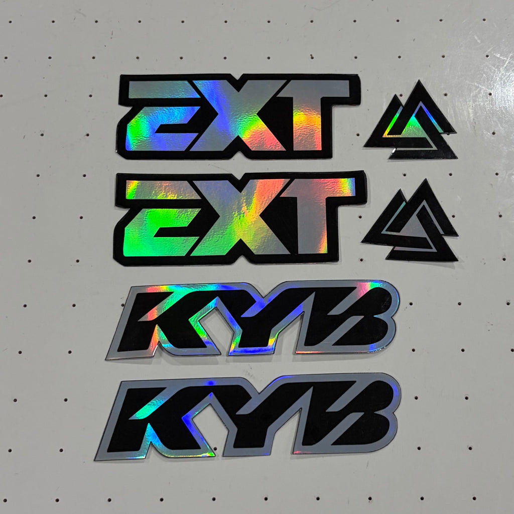 A set of four custom decal stickers featuring '2XT' and 'KYB' logos with a holographic rainbow sheen, displayed on a white pegboard workbench.