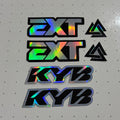 A set of four custom decal stickers featuring '2XT' and 'KYB' logos with a holographic rainbow sheen, displayed on a white pegboard workbench.