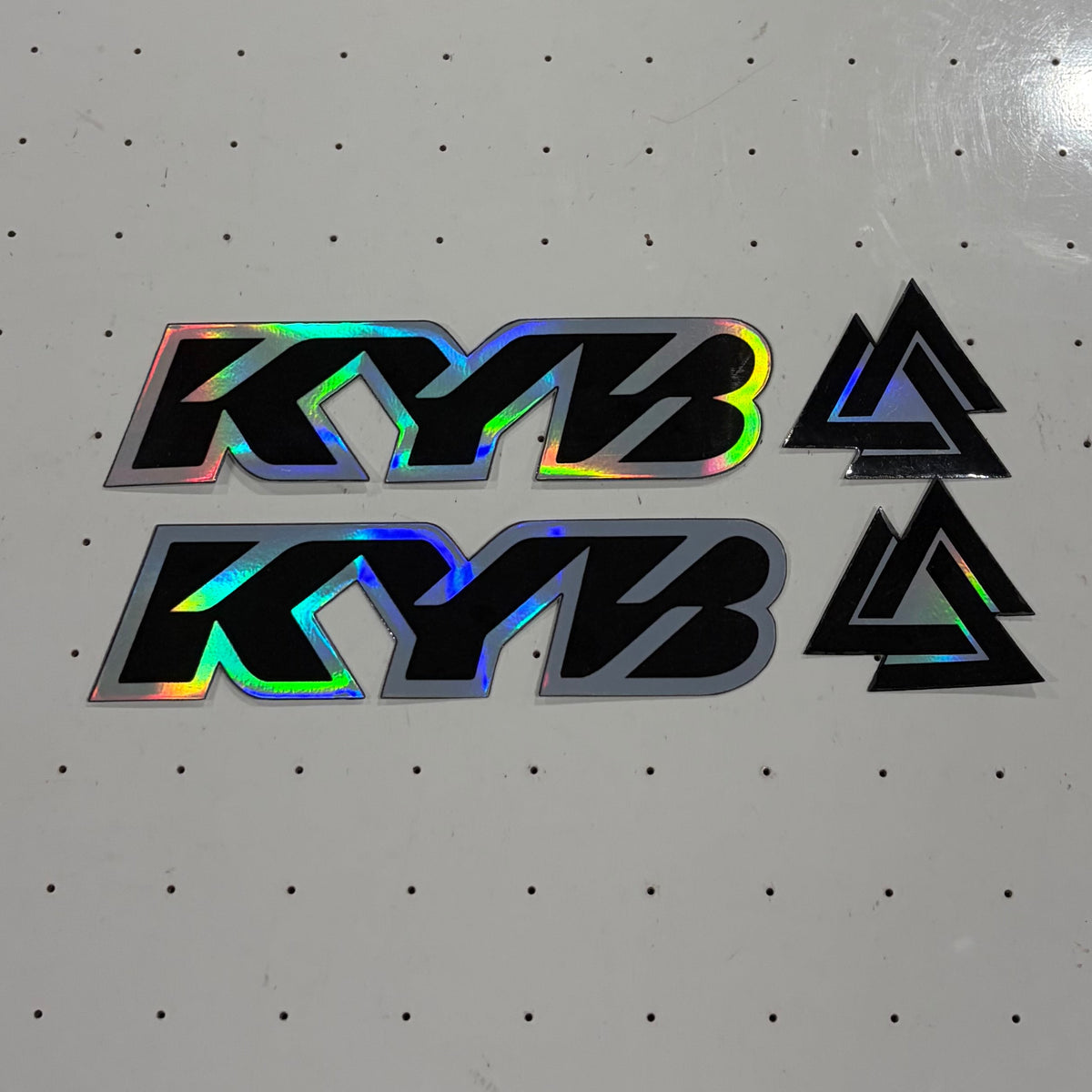 A set of holographic stickers featuring the text 'KYB' in black with a rainbow iridescent sheen, alongside black triangular logos, displayed on a white pegboard table.