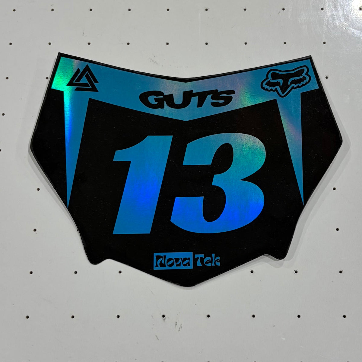 A motocross number plate decal with the number 13 in blue and green iridescent foil on a black background, featuring Fox and Reebok logos, placed on a white pegboard.