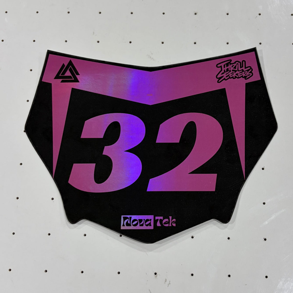 A motocross number plate sticker with the number 32 in a pink-to-purple gradient on a black background, displayed on a white pegboard.