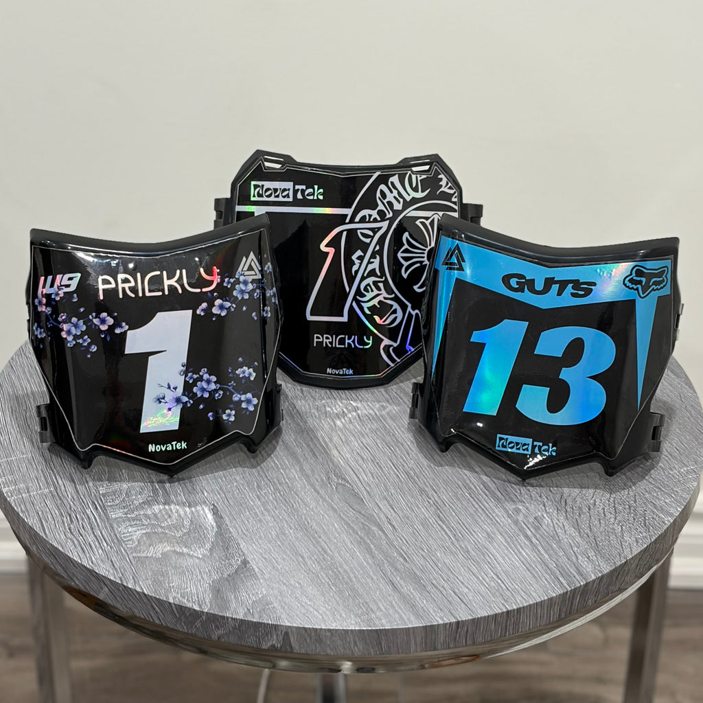 Three black motocross-style face shields with racing graphics and numbers are displayed on a round gray table against a plain indoor wall.