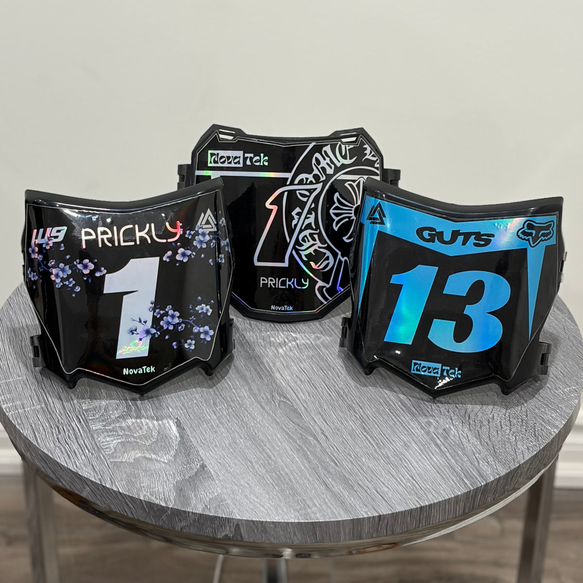 Three black motocross-style face shields with racing graphics and numbers are displayed on a round gray table against a plain indoor wall.