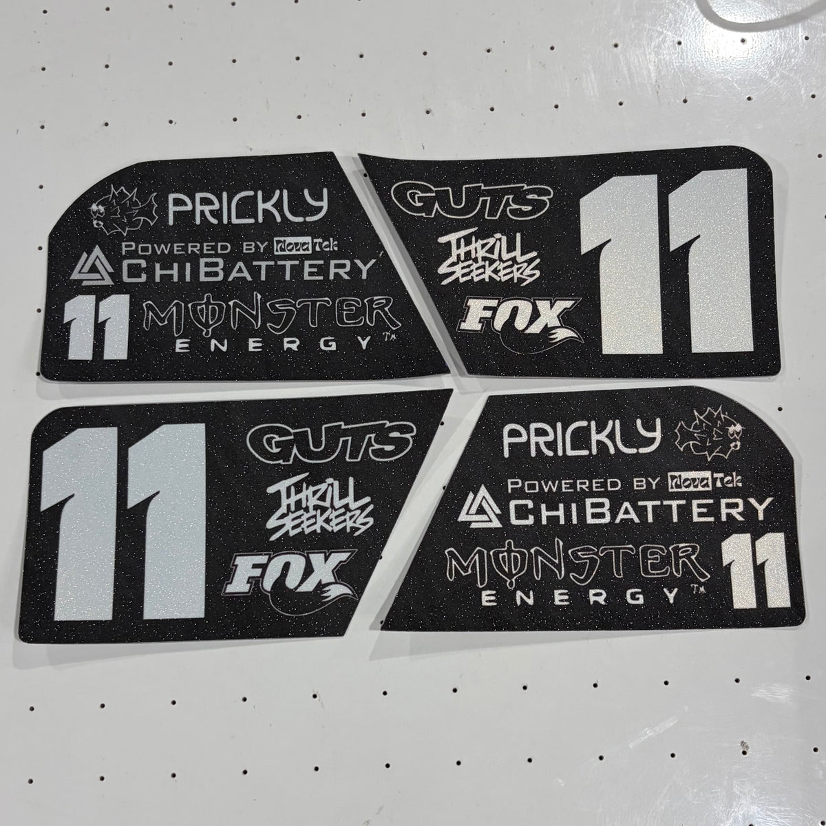 Four black foam decals with white and silver graphics for a racing livery, featuring the number 11 and sponsor logos like Prickly, ChiBattery, Guts, Thrill Seekers, and Fox, arranged on a white pegboard background.