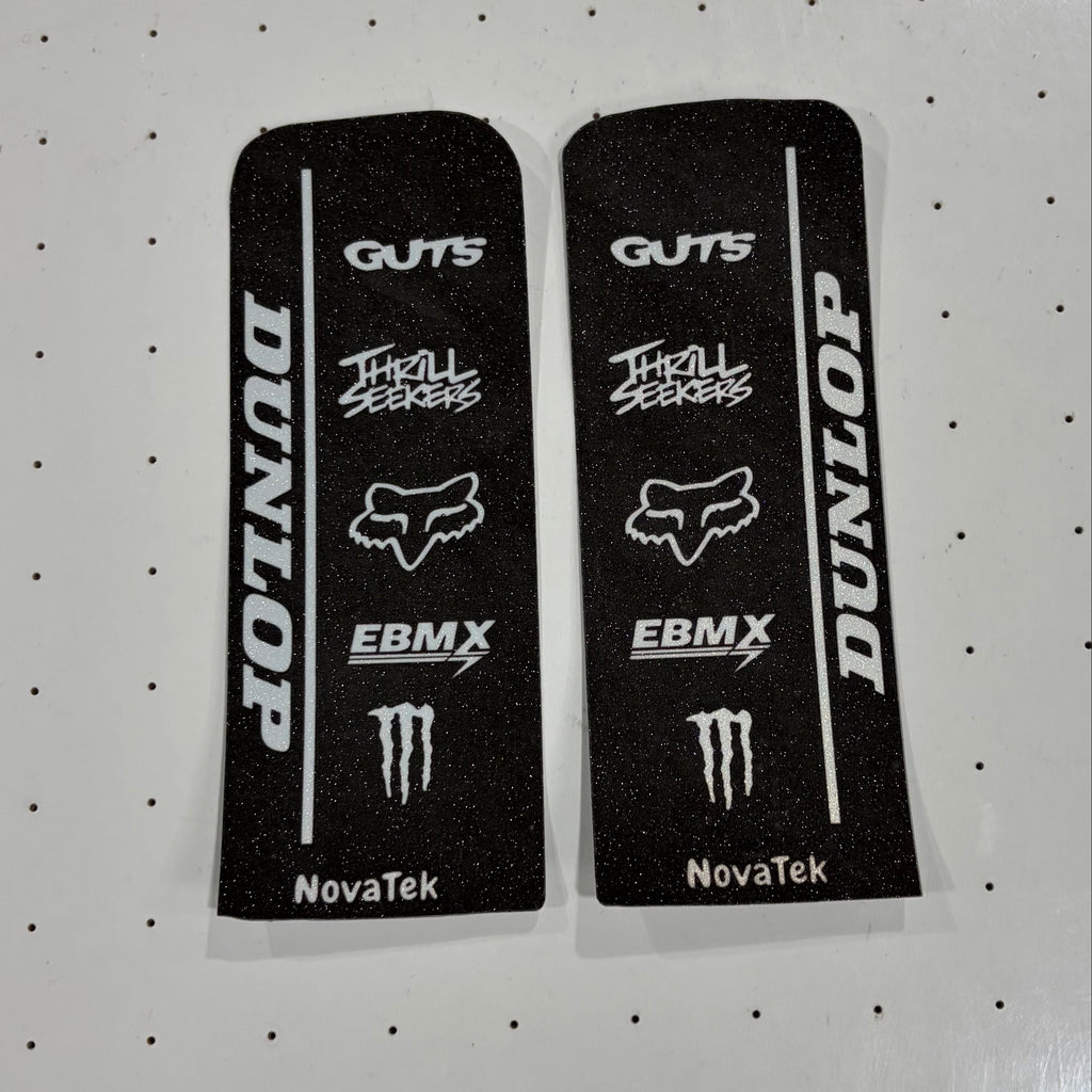 Two black rectangular decals with white Dunlop, Fox, EBMX, and Monster Energy logos, displayed side-by-side on a white pegboard.