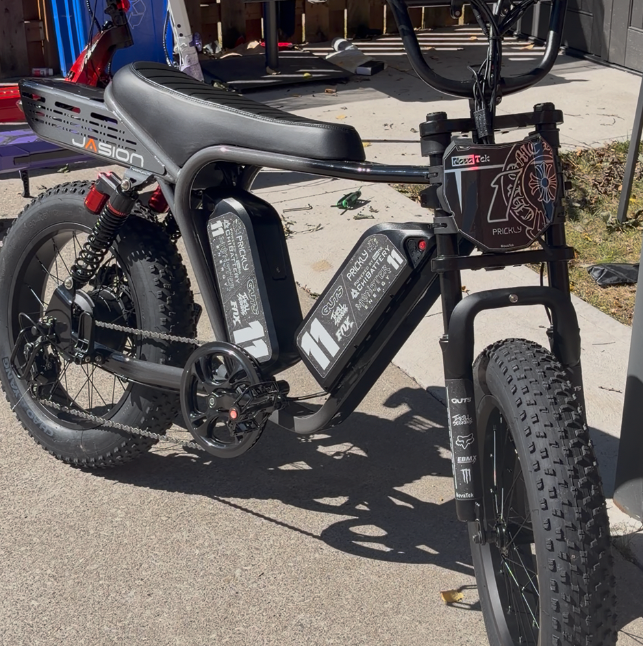 A black fat-tire electric bicycle parked on a concrete driveway in front of a wooden fence and gray shed, with a small basket on the front and a large battery mounted under the seat.