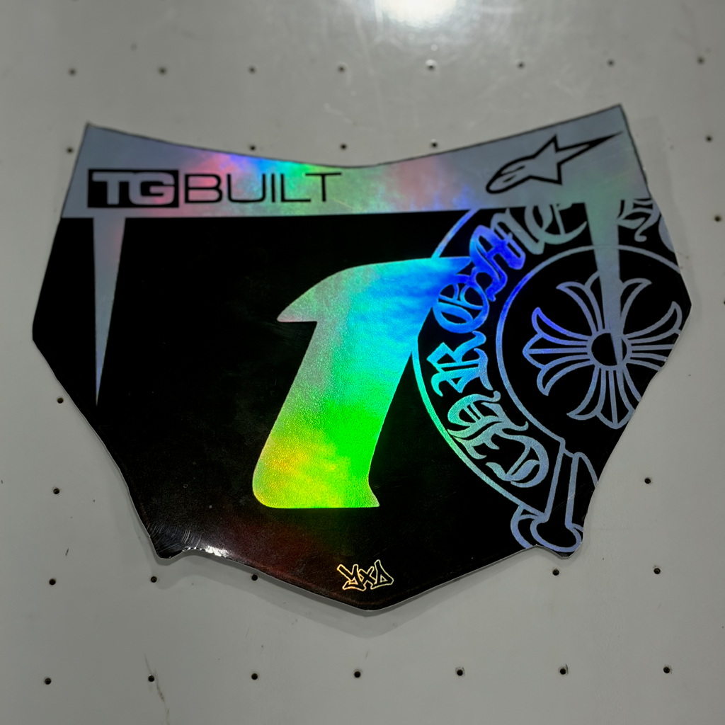 A black motocutz plate with a highly reflective, holographic silver finish, featuring a large holographic number '1' on the front, 'TBUILT' branding, and a blue graphic on the side, displayed on a white pegboard.