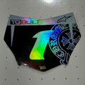 A black motocutz plate with a highly reflective, holographic silver finish, featuring a large holographic number '1' on the front, 'TBUILT' branding, and a blue graphic on the side, displayed on a white pegboard.