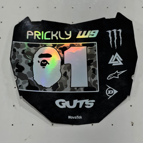 A black motorcycle number plate skin or decal with various sponsor logos, including A Bathing Ape, Monster Energy, and Alpinestars, displayed on a white pegboard.