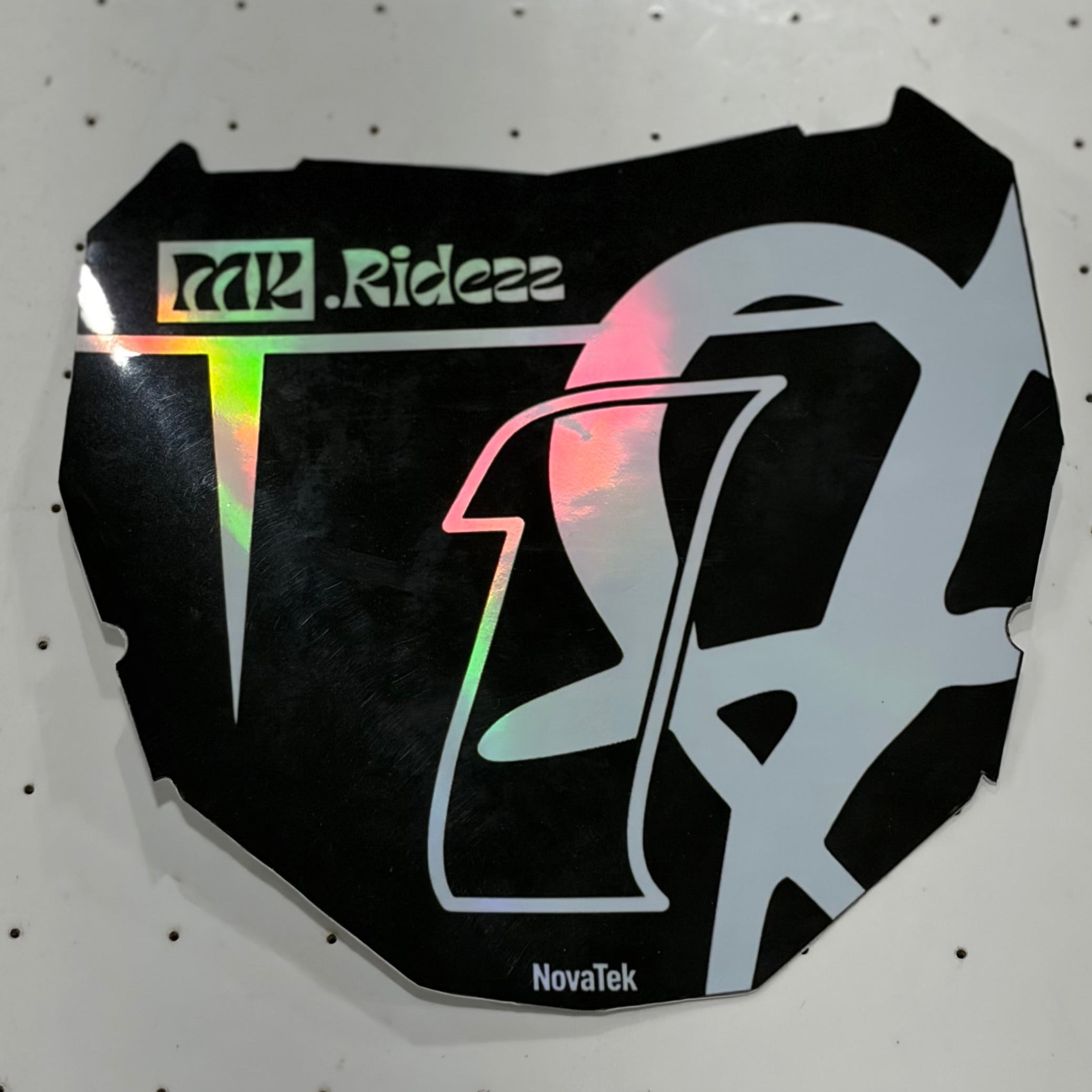 A black custom motorcycle number decal sticker with holographic silver graphics and 'Ride24' branding, displayed on a white pegboard background.