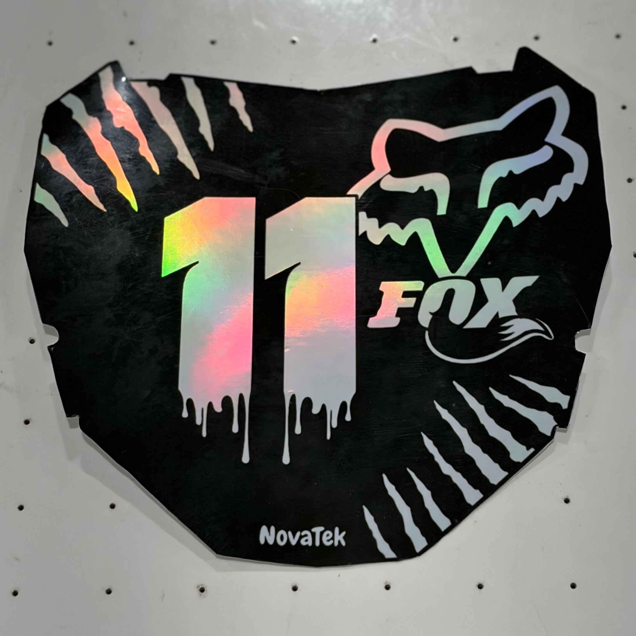 A black motorcycle number plate skin or decal with holographic silver '11' and white Fox Racing graphics, displayed on a white pegboard.