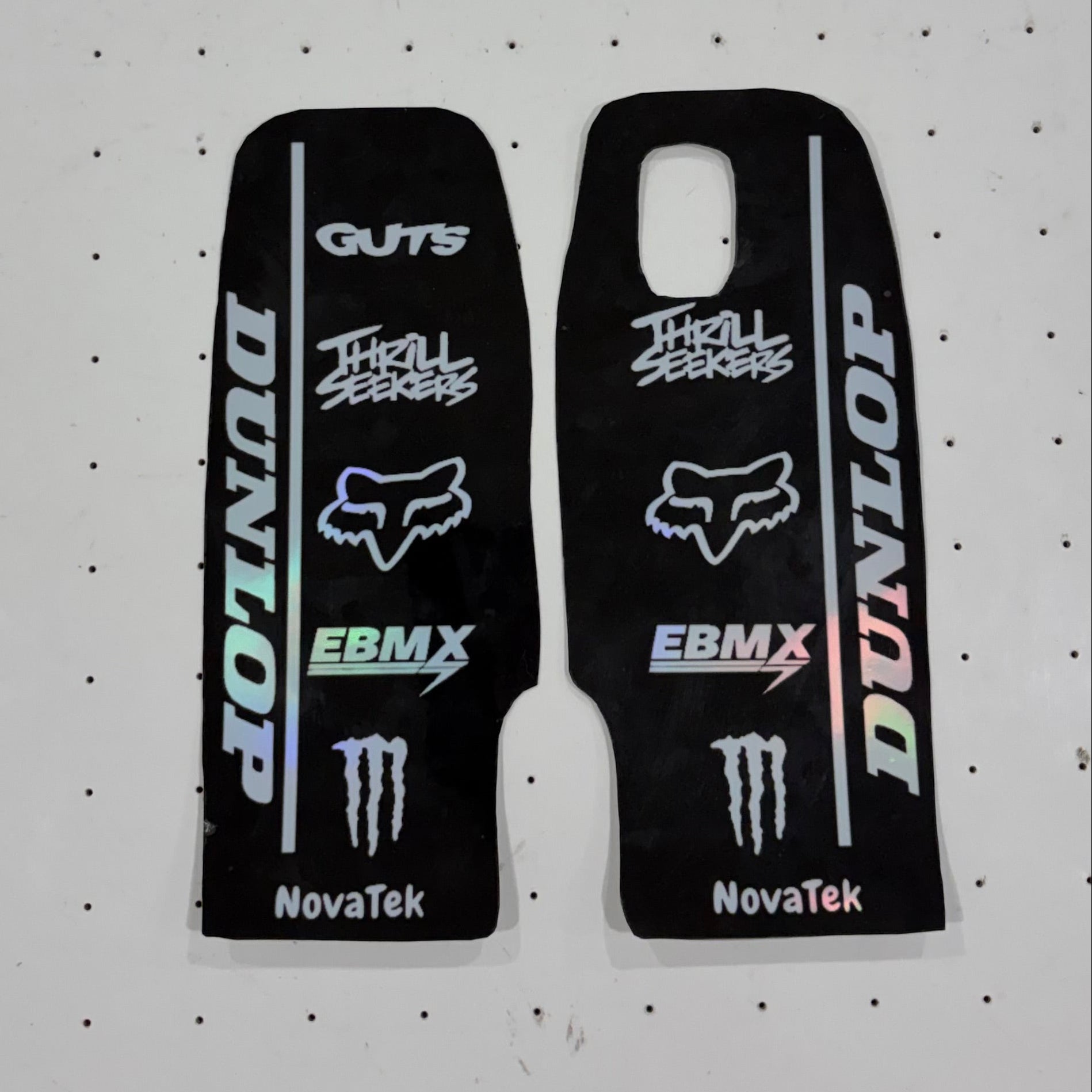 Two black motocross stickers with various sponsor decals, including Dunlop, Fox Racing, EBM X, Monster Energy, and NovaTek, displayed on a white pegboard background.