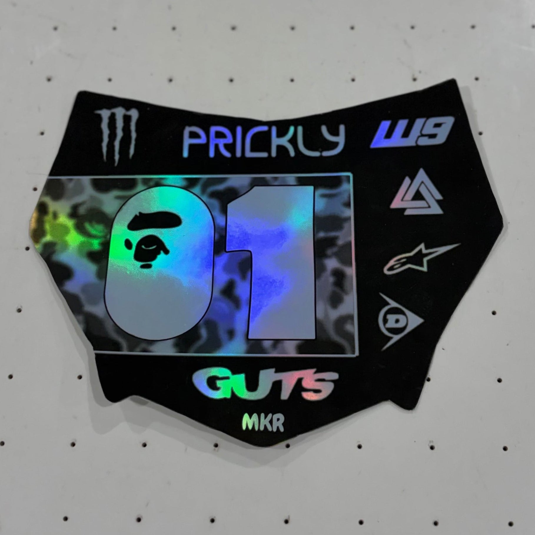 A black die-cut sticker featuring a holographic silver '01' with a leopard print background, placed on a white perforated surface. The sticker includes various brand logos such as Monster Energy, Prickly, W9, A Bathing Ape, Alpinestars, and Guts MKR.