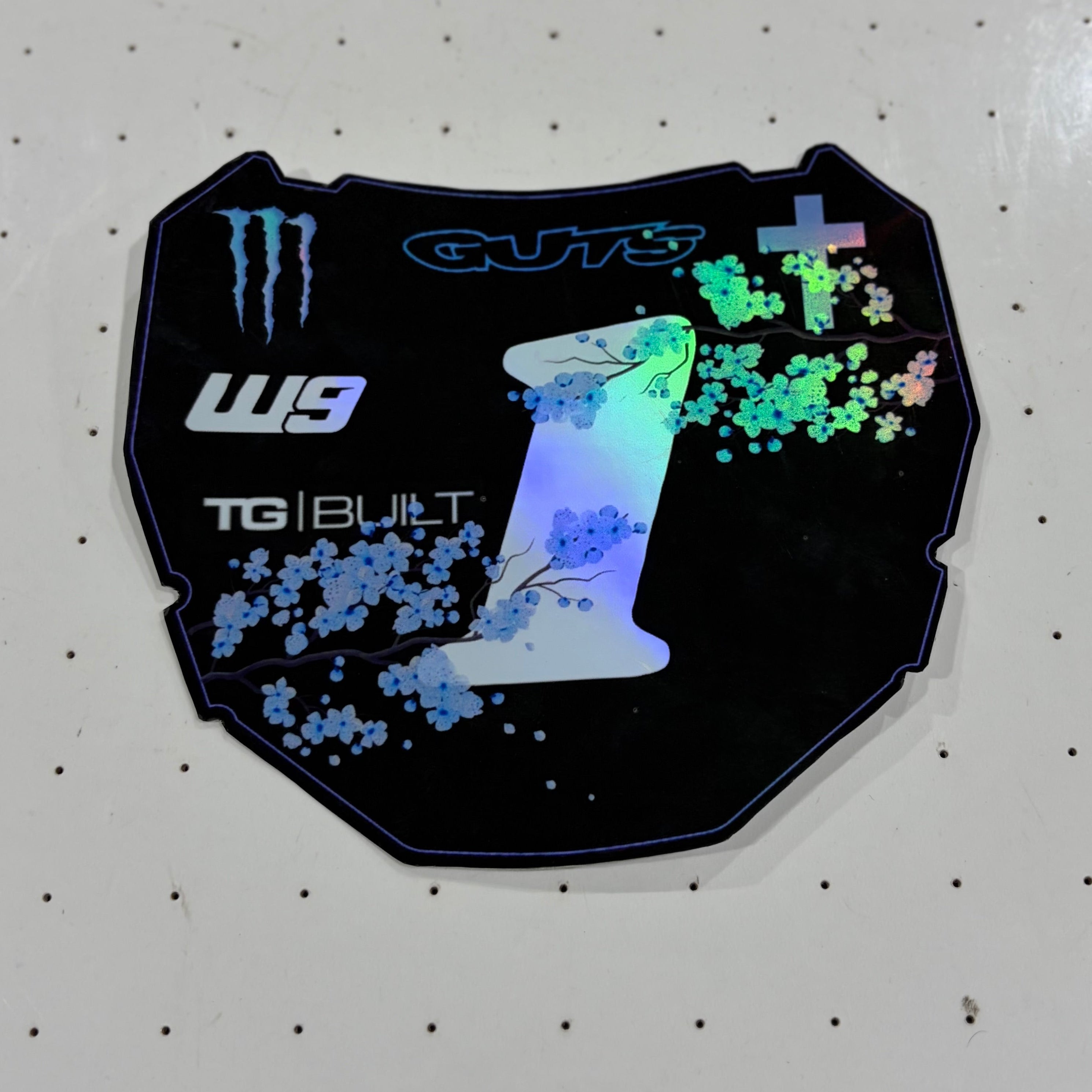 A black die-cut sticker featuring a stylized white number 1 with blue and green holographic splatter graphics, alongside Monster Energy and W9 branding, displayed on a white pegboard background.