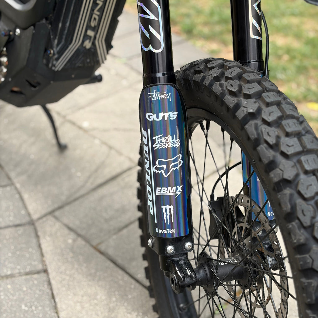 Close-up of a black ebike front fork with multiple sponsor logos including GUTS, Fox, and Monster Energy, mounted on a knobby tire on a paved outdoor path.