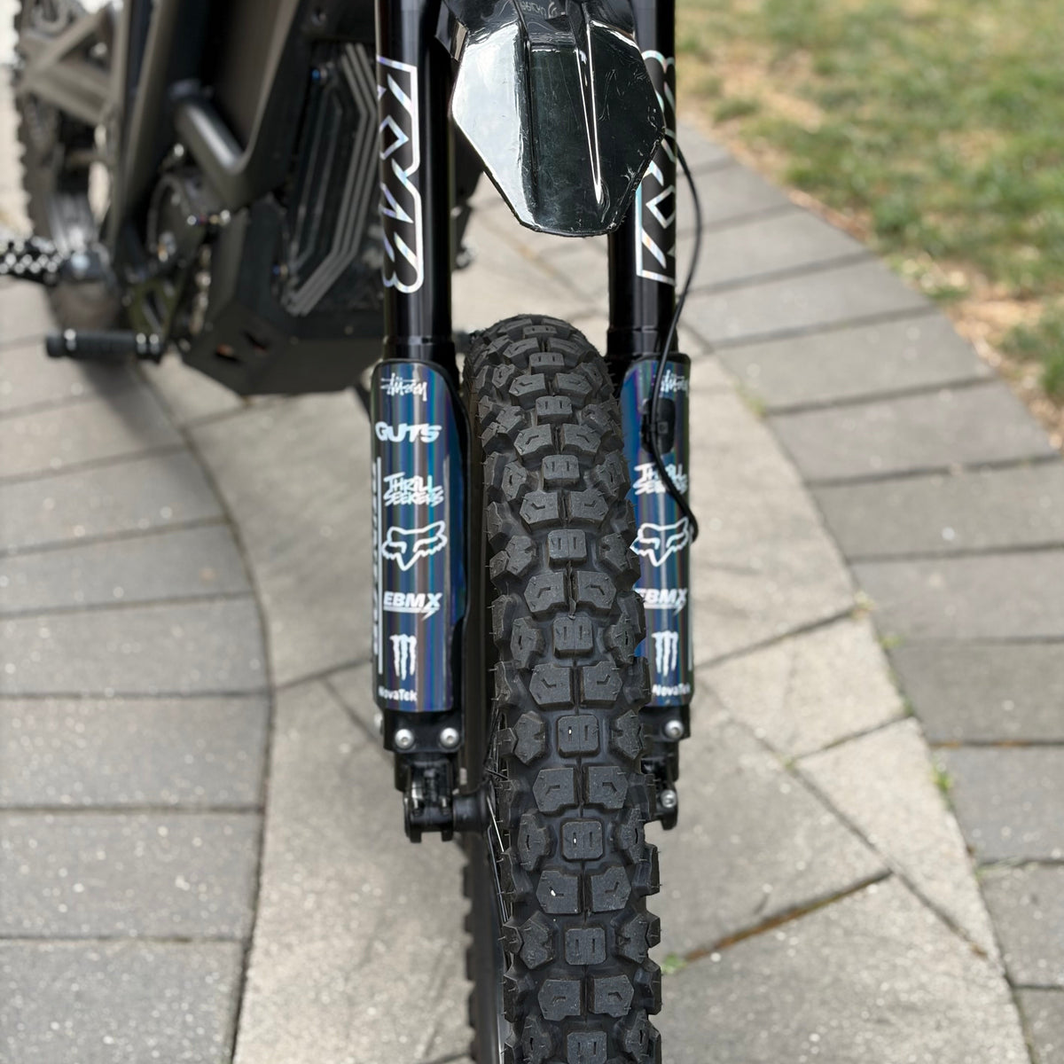 Close-up of a black off-road motorcycle or dirt bike front end, showing a knobby tire, black fork shrouds, blue Fox suspension components, and a black handlebar cage with graphics, photographed outdoors on a paved path.