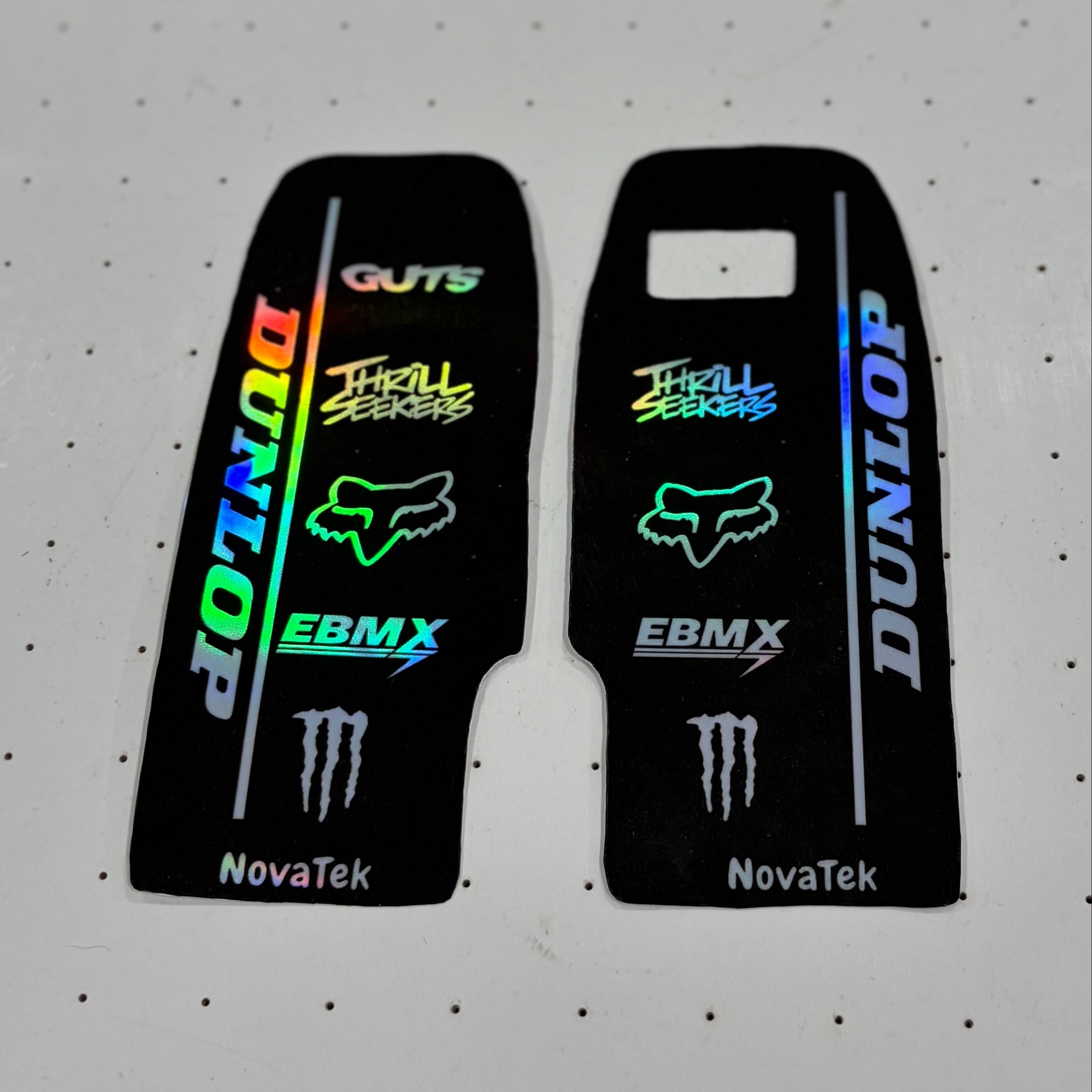 Two black motorcycle tank decal stickers with various sponsor logos, including Dunlop, Fox Racing, EBM X, and Monster Energy, displayed on a white pegboard background.