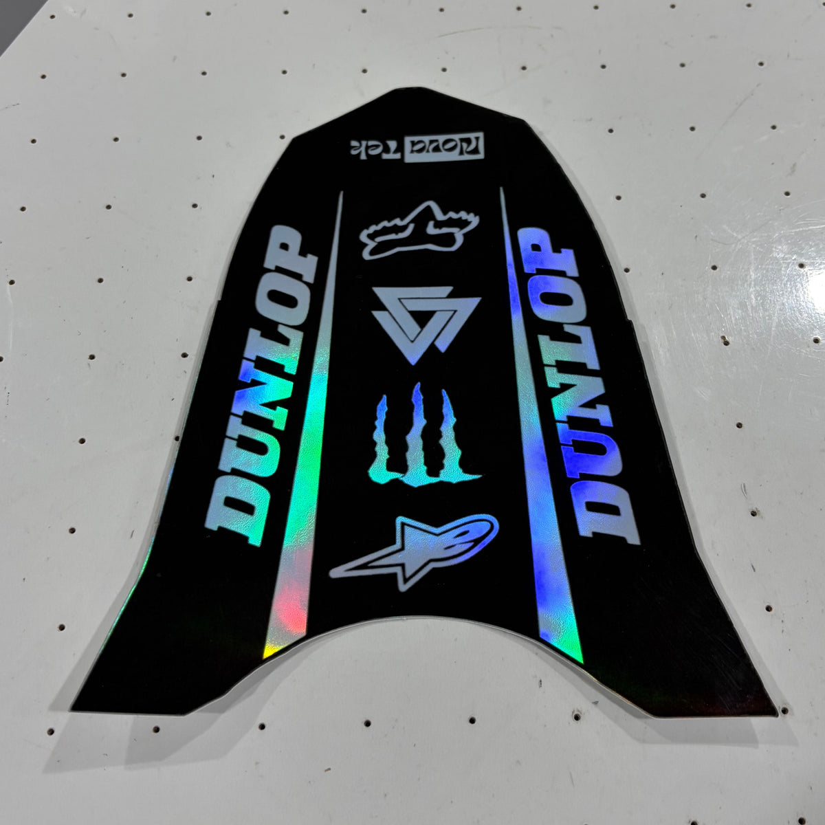 A black motorcycle or electric dirt bike decal sticker set featuring Dunlop branding, Alpinestars logos, and holographic rainbow accents, displayed on a white pegboard surface.