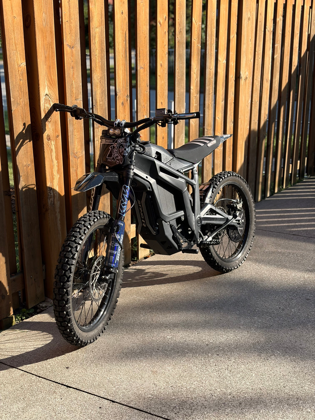 A black electric dirt bike with a blue front fork is parked on concrete next to a wooden fence.
