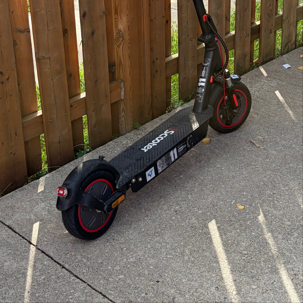 A black and red electric scooter with branding leans against a tall wooden fence on a concrete patio.