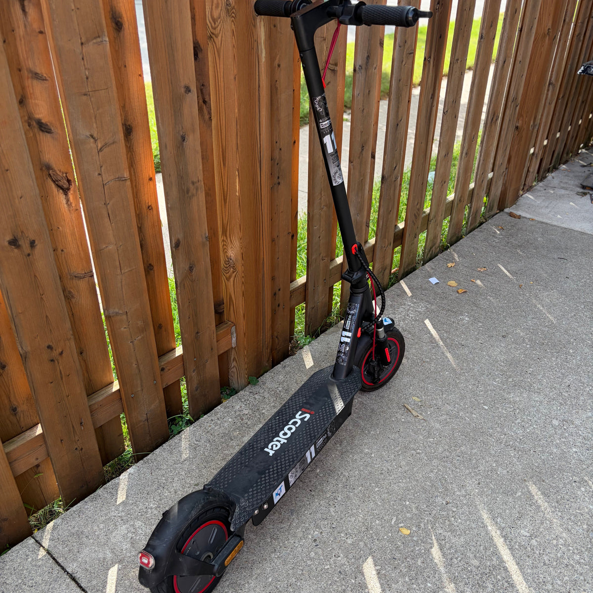 A black and red electric scooter parked on a concrete path next to a wooden fence.