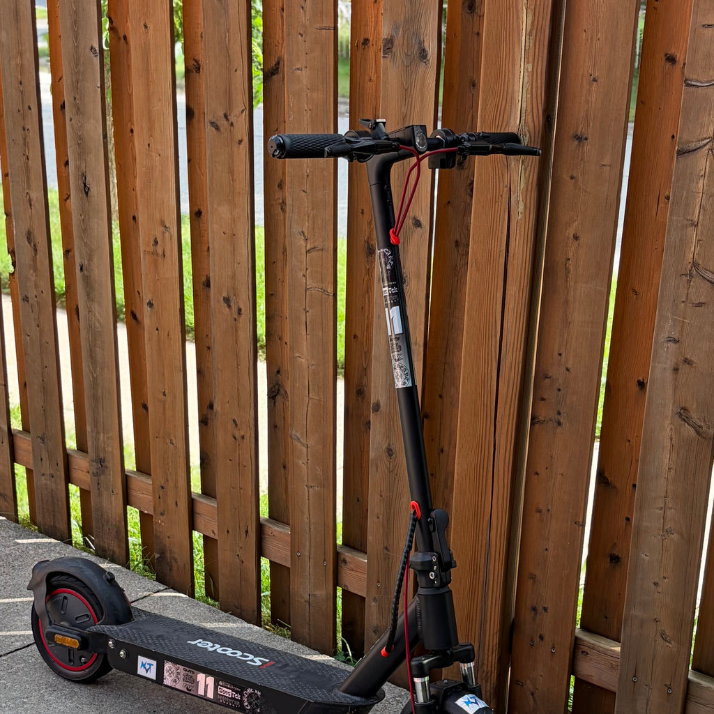 A black electric scooter with red accents is parked on a concrete path next to a tall wooden fence.