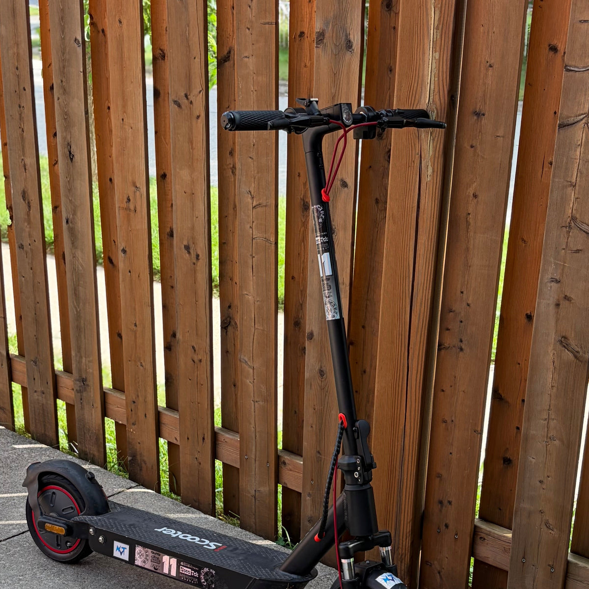 A black electric scooter with red accents is parked on a concrete path next to a tall wooden fence.