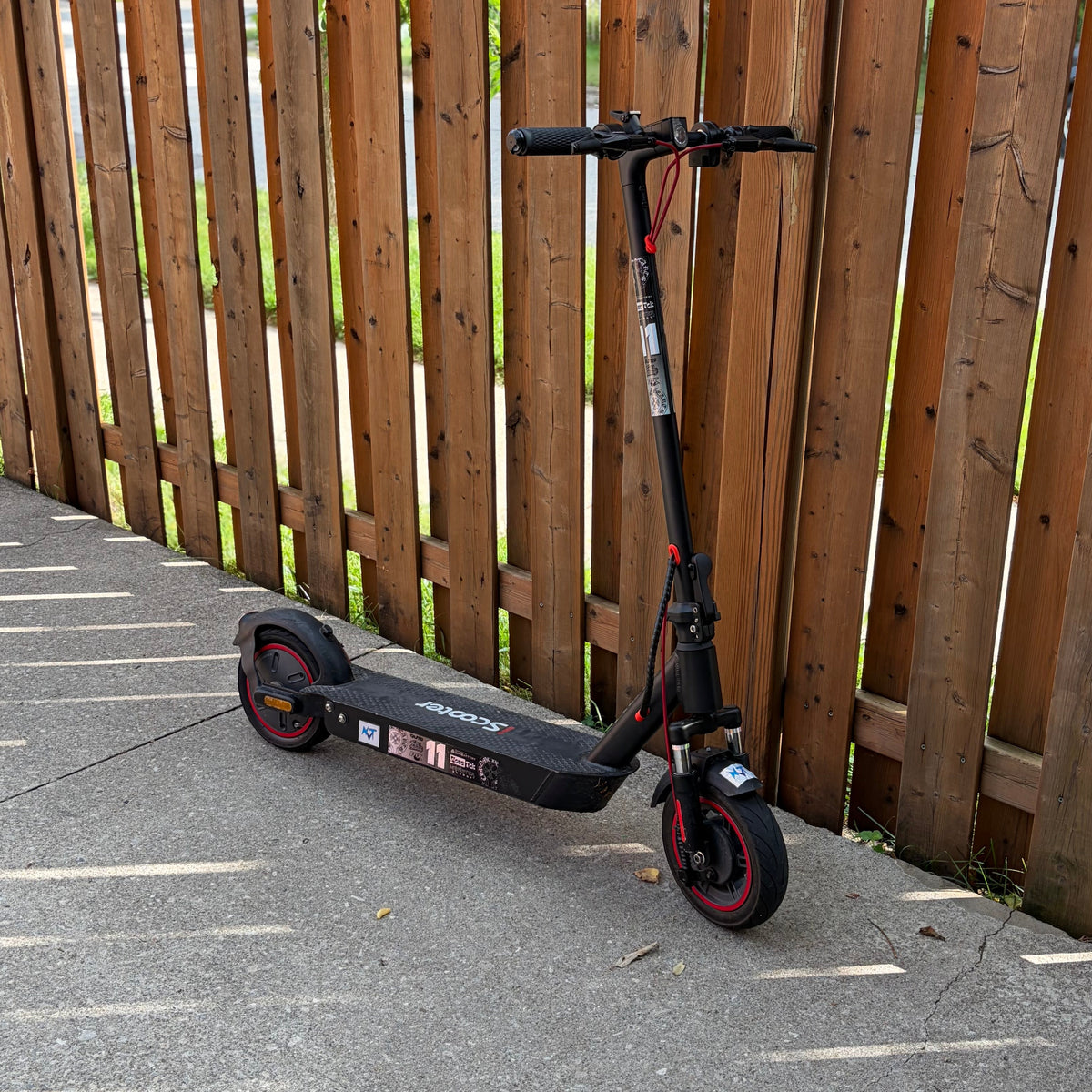 A black electric scooter with red accents is parked on a concrete path next to a tall wooden fence.