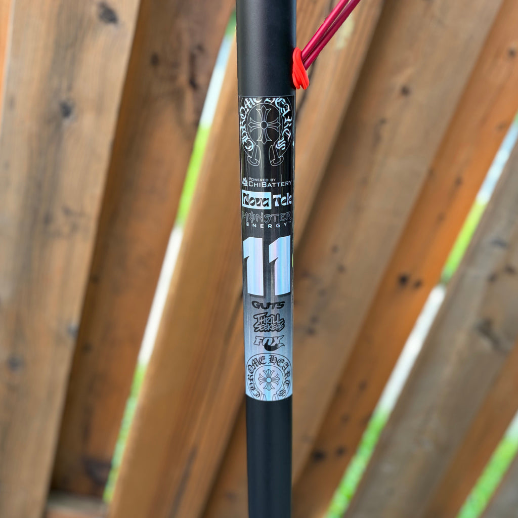 Close-up of a black bicycle frame tube with a silver and black Cannondale 11 sticker, featuring a red cable loop against a wooden fence background.
