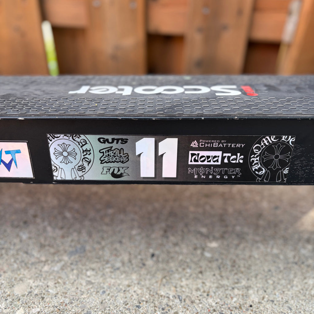 A close-up of a black scooter deck featuring various stickers, including 'Guts', 'Road Tech', and 'Chibattery', with the number '11' printed on the side. The background shows a blurred wooden fence and concrete ground.