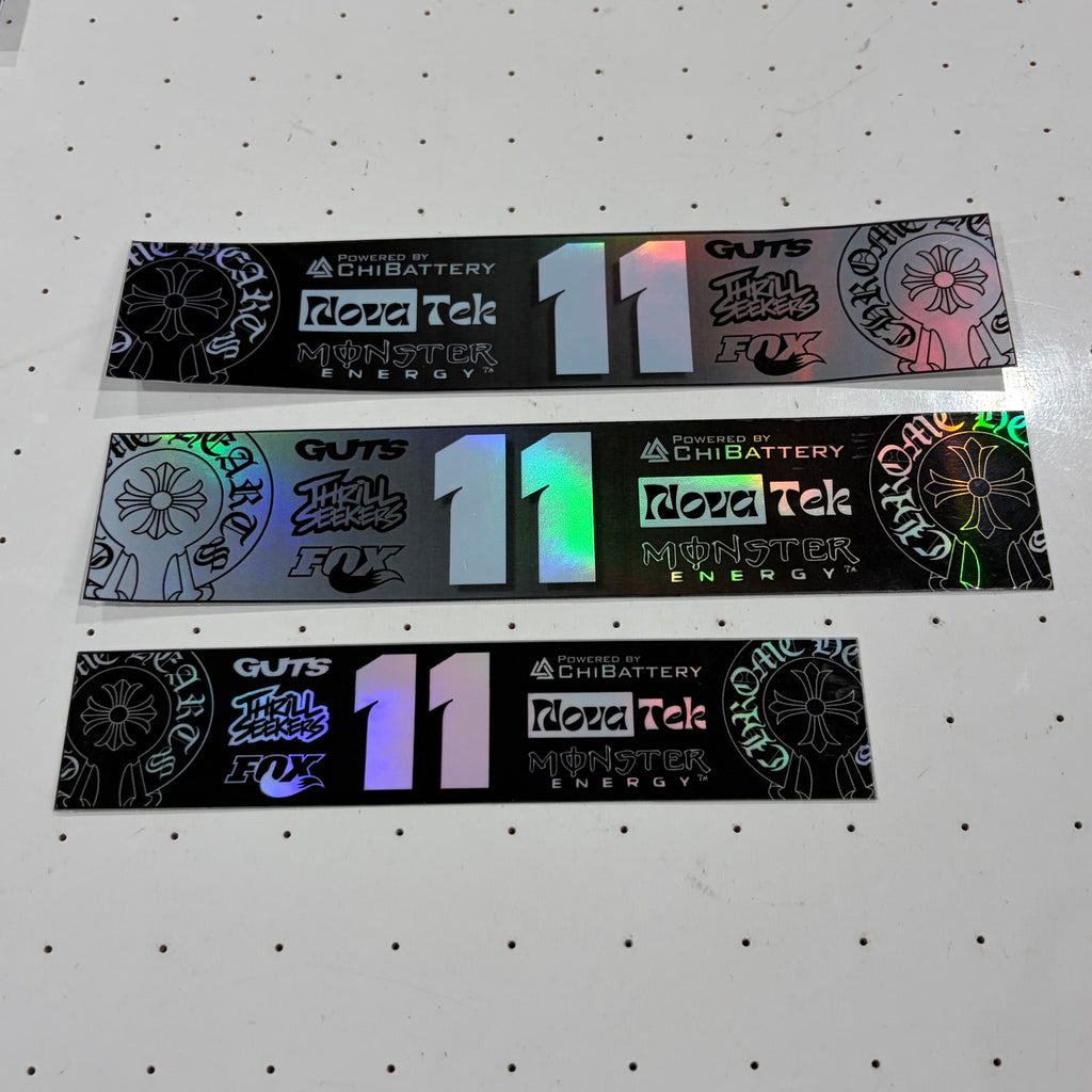 Three rectangular black stickers with holographic and multicolor printed graphics, laid horizontally on a white pegboard background. The stickers feature the text 'GUTS', 'Chibattery', 'Royal Tek Monster Energy', and a large number '11'.