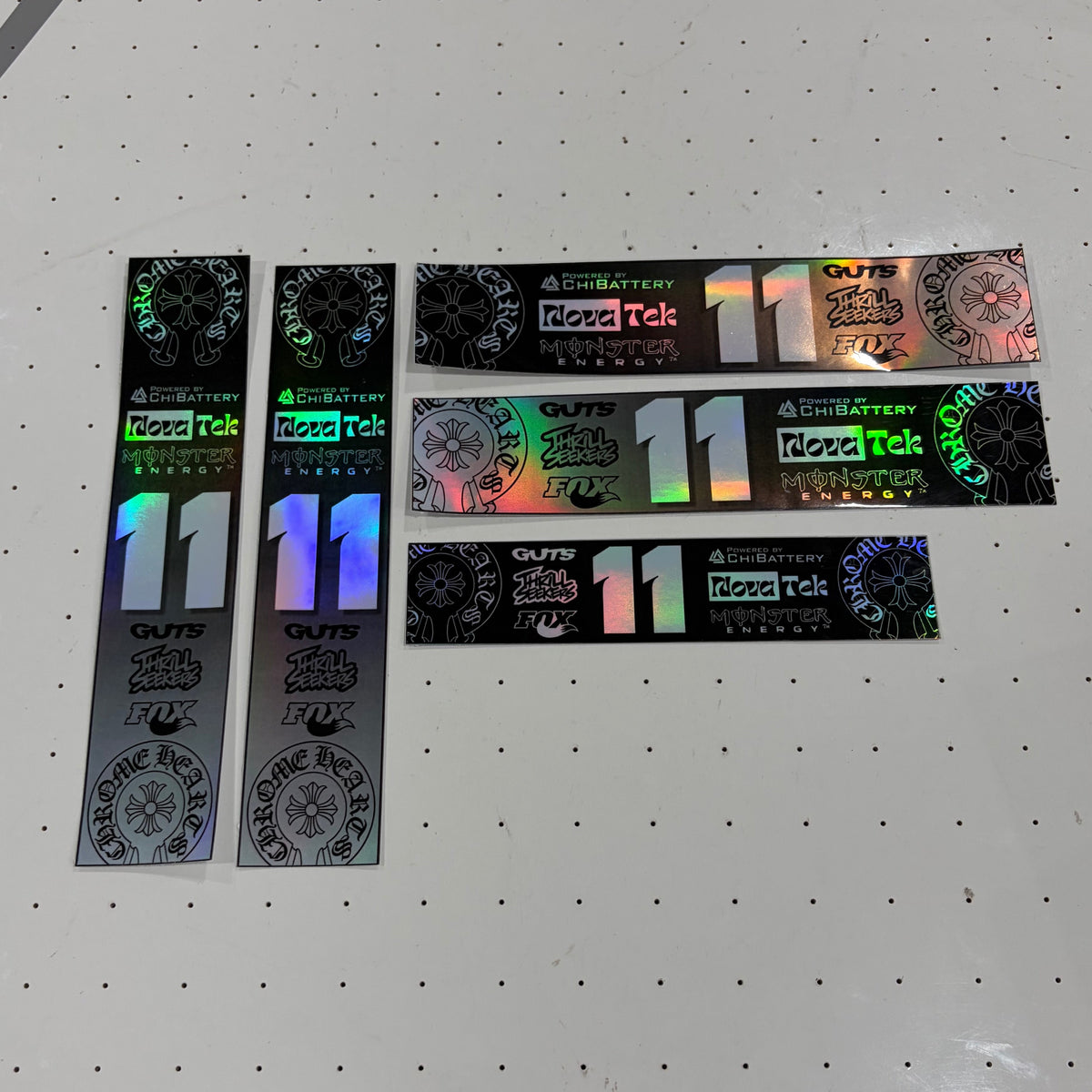 Several long rectangular stickers with holographic iridescent printing and the number 11 are arranged on a white pegboard surface.