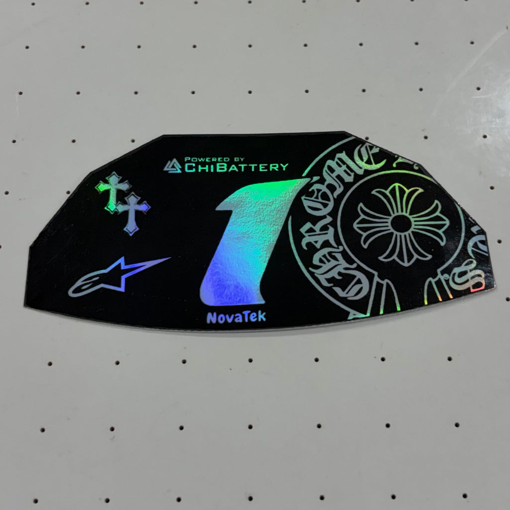 A black die-cut sticker with holographic rainbow effects and Alpinestars branding, placed on a white perforated surface.