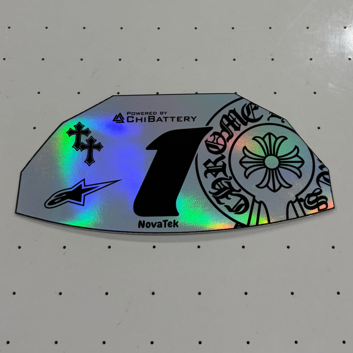 A close-up of a custom motorcycle number decal sticker with holographic silver foil, featuring a large black number 1 and various brand logos including NovaTek, EmsaBattery, and Alpinestars, displayed on a white pegboard.