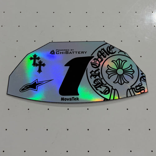A close-up of a custom motorcycle number decal sticker with holographic silver foil, featuring a large black number 1 and various brand logos including NovaTek, EmsaBattery, and Alpinestars, displayed on a white pegboard.