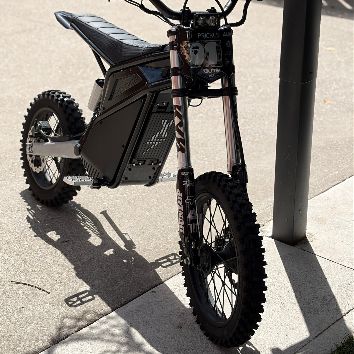 A black and silver electric dirt bike with knobby tires, parked on a concrete surface outdoors next to a wooden fence and a metal pole.