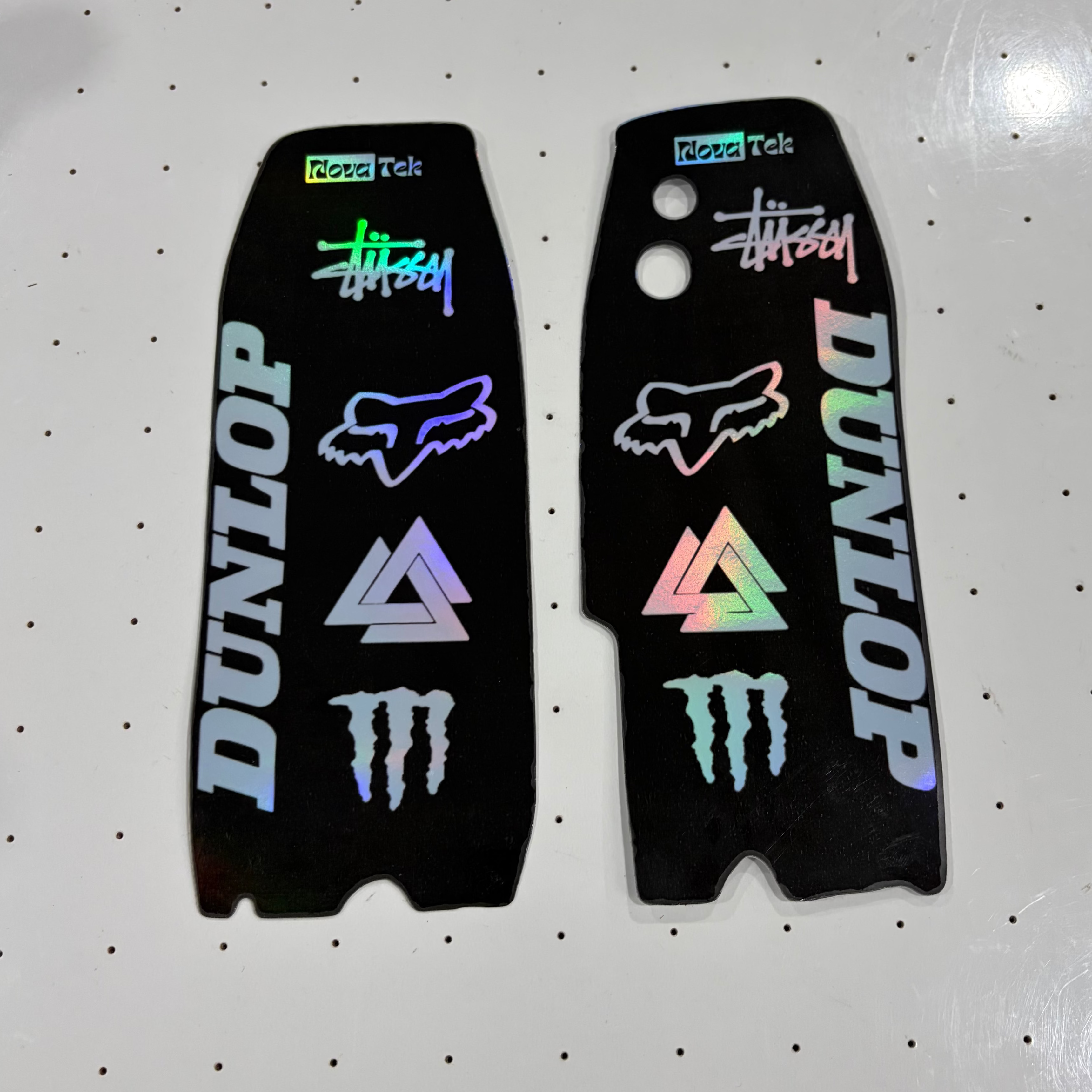Two black motorcycle tank decals with various sponsor logos including Dunlop, Fox, Monster Energy, and Stüssy, displayed on a white pegboard background.